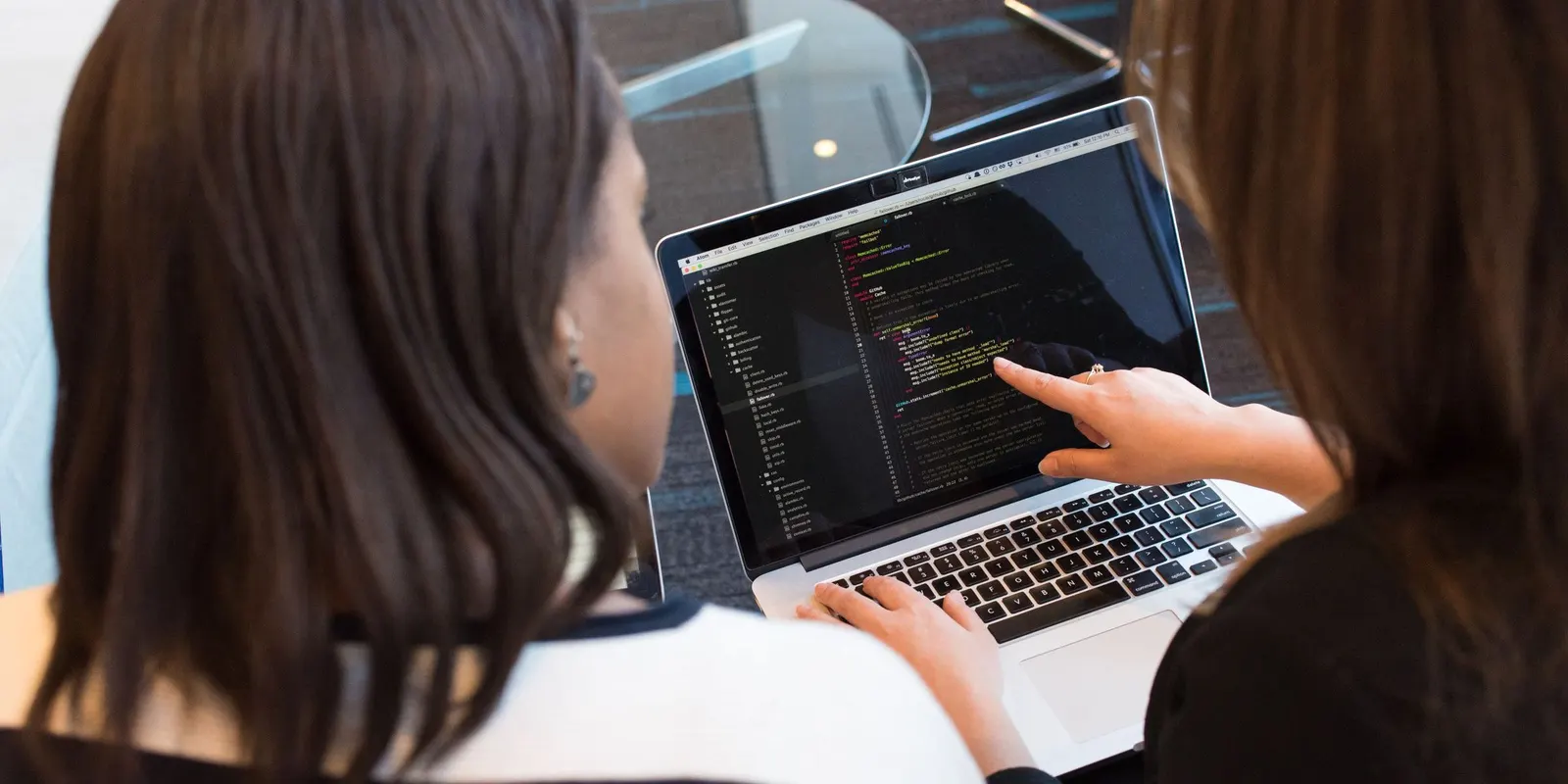 Zwei Entwicklerinnen prüfen gemeinsam Code am Laptop, eine zeigt auf eine Stelle im Editor